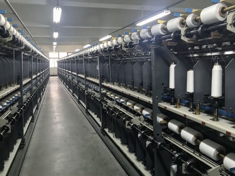 Textile factory production workshop with rows of industrial yarn spinning machines and textile manufacturing equipment.